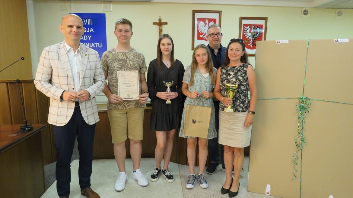 Uczestnicy uroczystości wręczenia nagród stoją razem w sali, trzymając puchary i dyplomy.