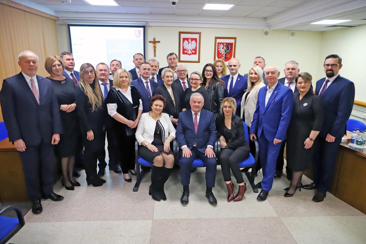 Zdjęcie przedstawia grupę osób pozujących do fotografii w sali z ikonografią narodową, wśród nich znajduje się kilku mężczyzn i kobiet w formalnych strojach.