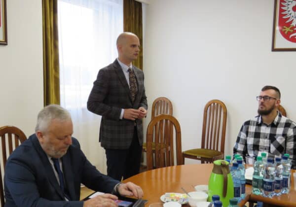 Mężczyzna w garniturze przemawia podczas spotkania w sali konferencyjnej, a wokół niego siedzą uczestnicy przy stole.