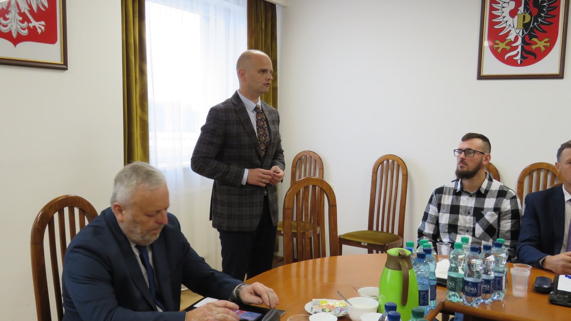 Mężczyzna w garniturze przemawia podczas spotkania w sali konferencyjnej, a wokół niego siedzą uczestnicy przy stole.