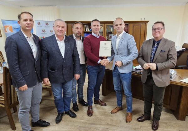 Na zdjęciu widać grupę sześciu mężczyzn, którzy stoją w biurze, a jeden z nich przekazuje dokument w rękach drugiego.