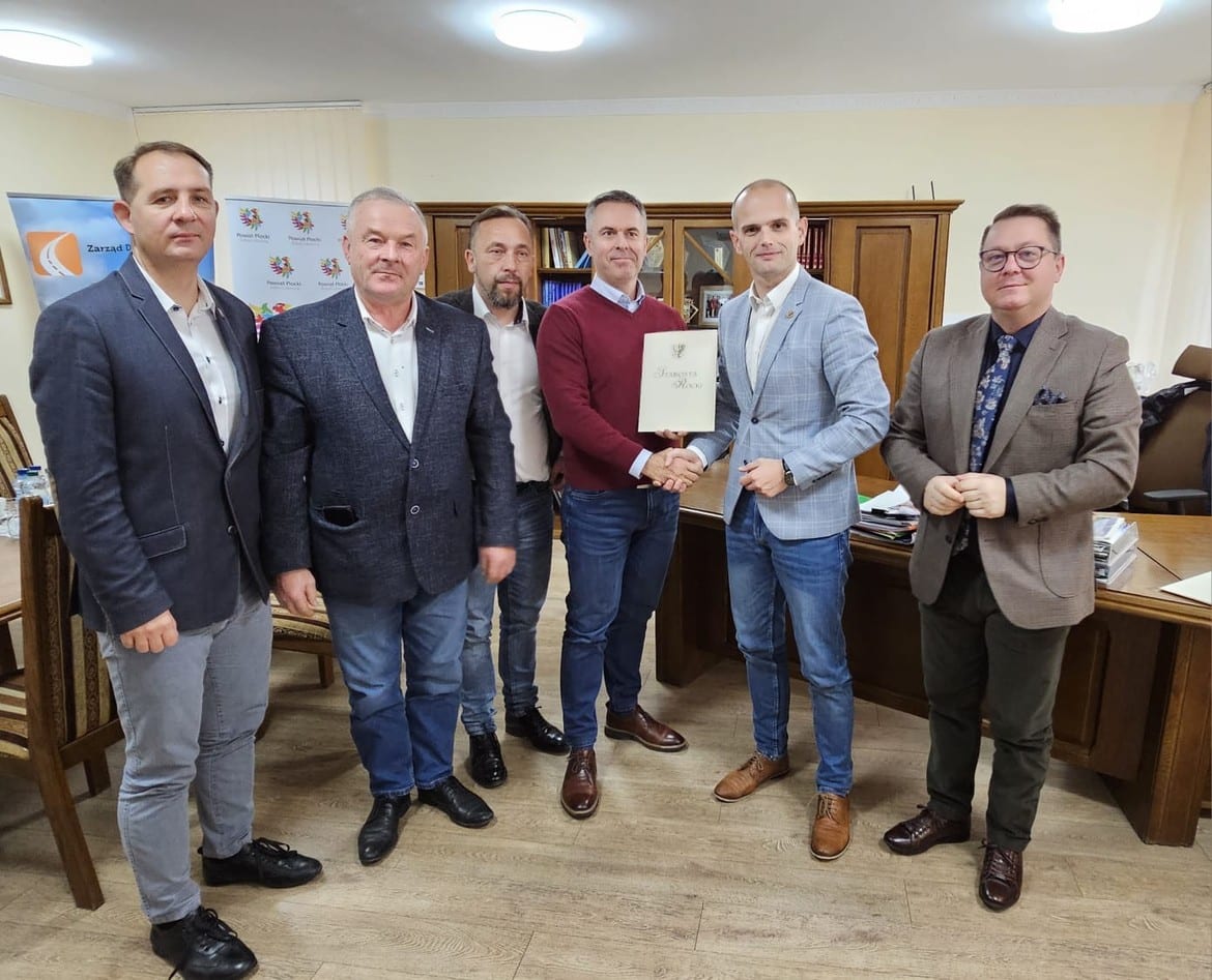 Na zdjęciu widać grupę sześciu mężczyzn, którzy stoją w biurze, a jeden z nich przekazuje dokument w rękach drugiego.