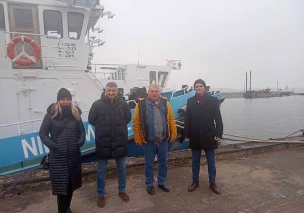 Na zdjęciu widoczna jest czwórka ludzi stojących na pomoście obok łodzi w mglistej scenerii portu.