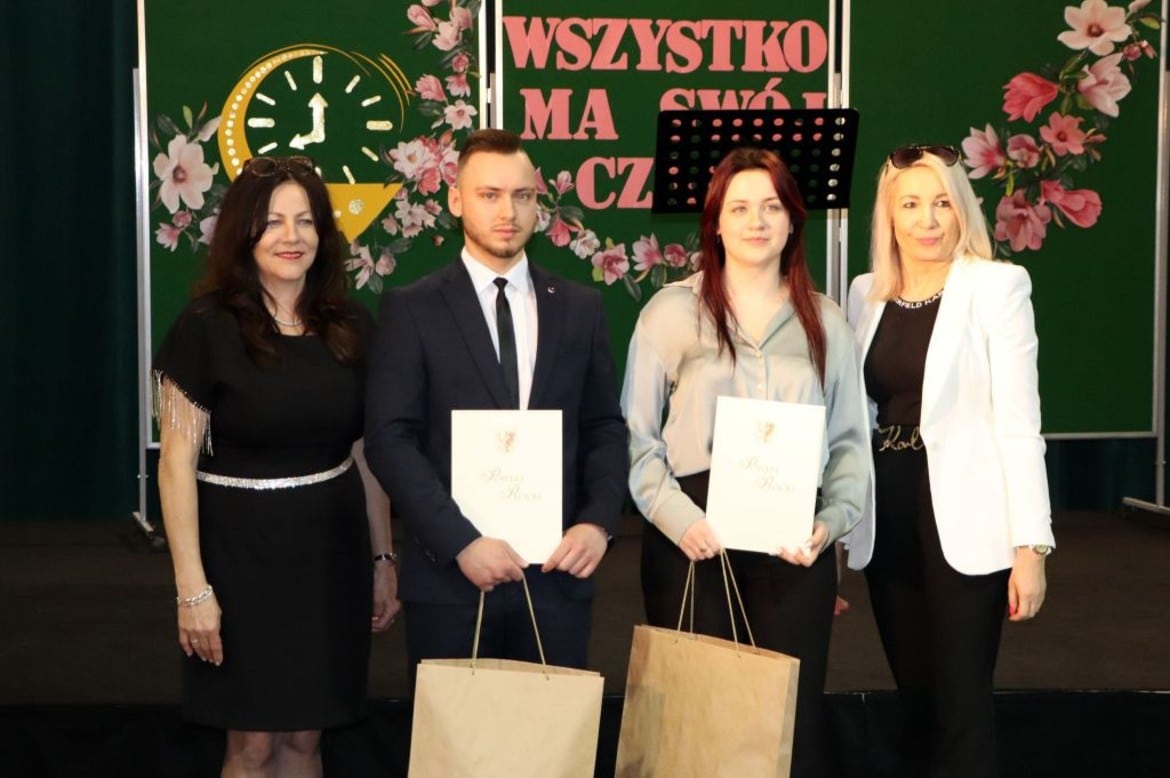 Na zdjęciu przedstawione są cztery osoby, które uczestniczą w uroczystości wręczenia dyplomów, stojąc na scenie przed kolorowym tłem z kwiatami i napisem „WSZYSTKO MA SWÓJ CZAS”.