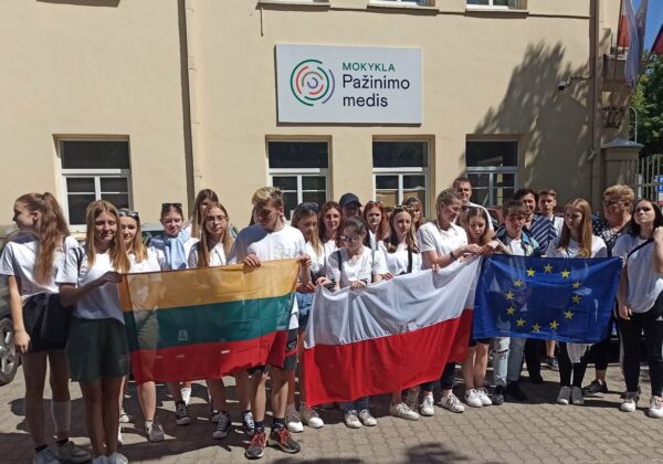 Grupa młodzieży stoi przed budynkiem szkoły, trzymając flagi Litwy, Polski oraz Unii Europejskiej.