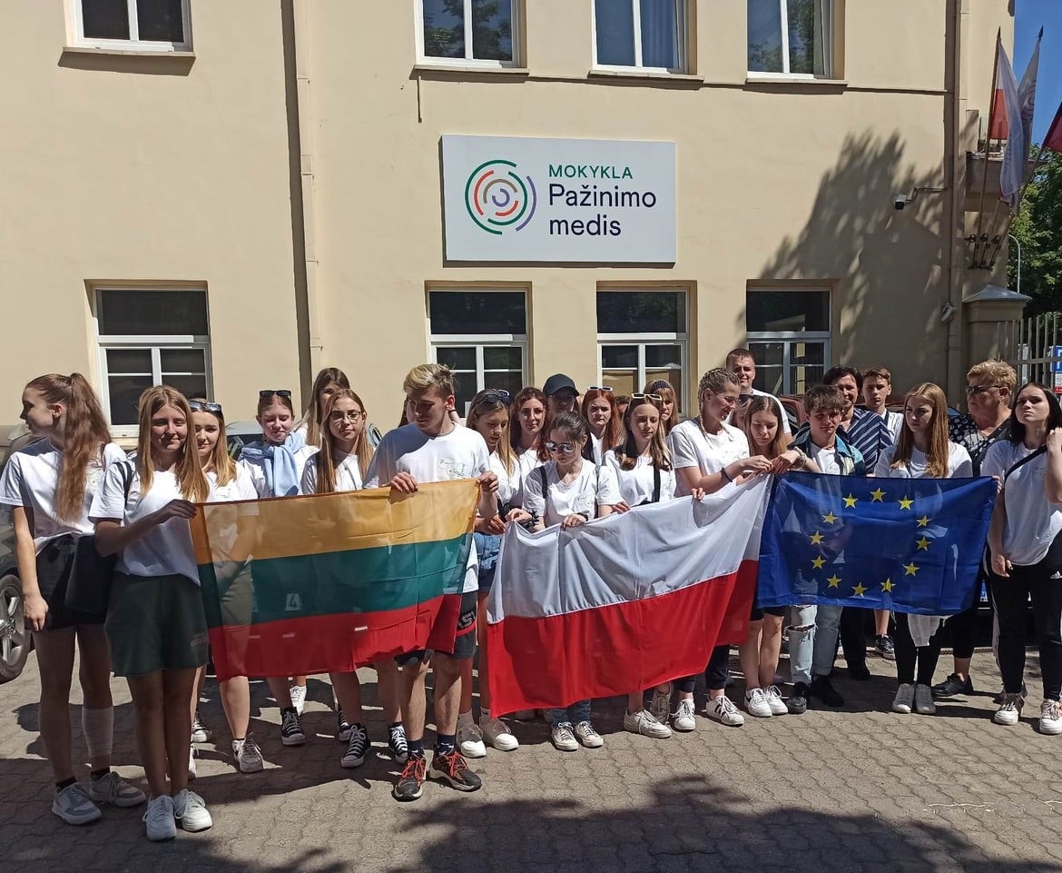 Grupa młodzieży stoi przed budynkiem szkoły, trzymając flagi Litwy, Polski oraz Unii Europejskiej.