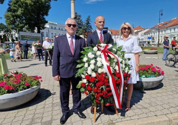 Na zdjęciu widoczna jest grupa ludzi trzymających wieniec z kwiatów, symbolizujący pamięć o ważnych wydarzeniach, na tle kwietników oraz budynków w słonecznym dniu.