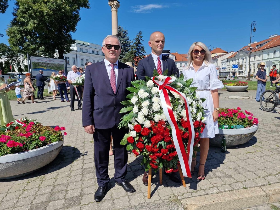 Na zdjęciu widoczna jest grupa ludzi trzymających wieniec z kwiatów, symbolizujący pamięć o ważnych wydarzeniach, na tle kwietników oraz budynków w słonecznym dniu.