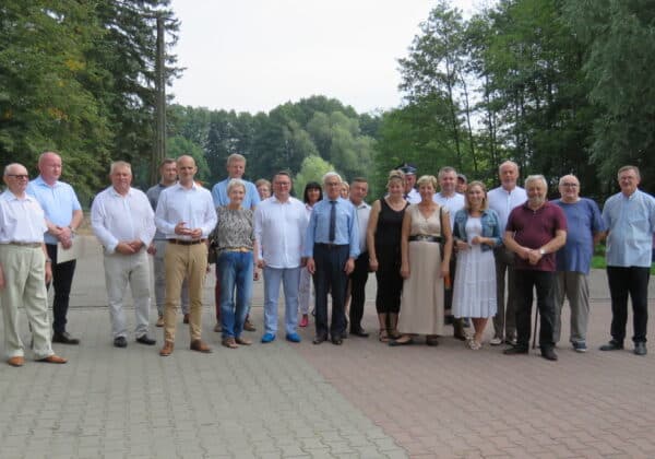 Na zdjęciu widać grupę osób stojących razem na świeżym powietrzu, w otoczeniu zieleni, w różnorodnych strojach, podczas spotkania lub wydarzenia.
