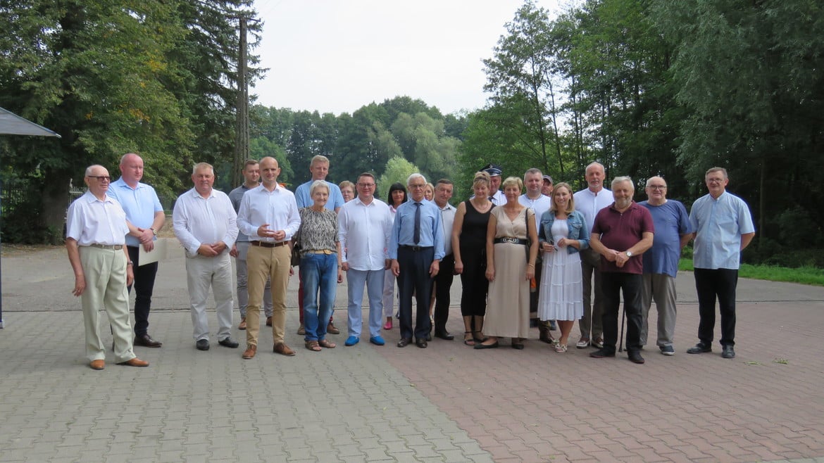 Na zdjęciu widać grupę osób stojących razem na świeżym powietrzu, w otoczeniu zieleni, w różnorodnych strojach, podczas spotkania lub wydarzenia.