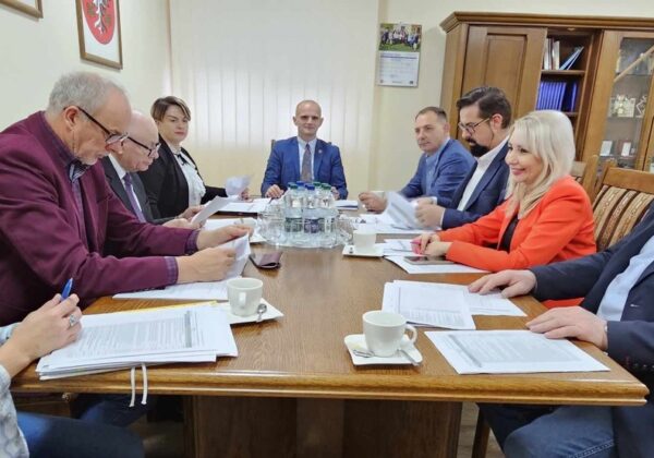 Na zdjęciu widać grupę osób podczas spotkania w sali konferencyjnej, siedzących przy stole z dokumentami oraz filiżankami.