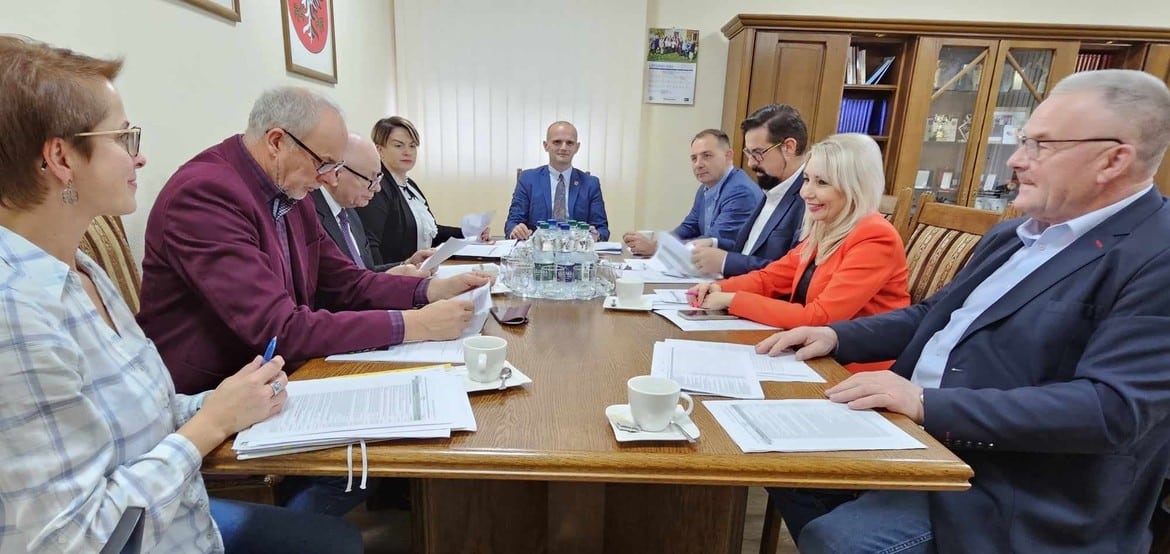 Na zdjęciu widać grupę osób podczas spotkania w sali konferencyjnej, siedzących przy stole z dokumentami oraz filiżankami.