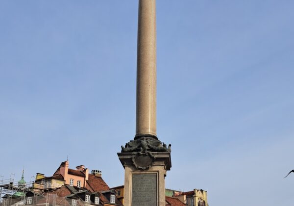 Grupa młodych ludzi stoi przed Kolumną Zygmunta w Warszawie, z widocznymi kamienicami w tle i błękitnym niebem.