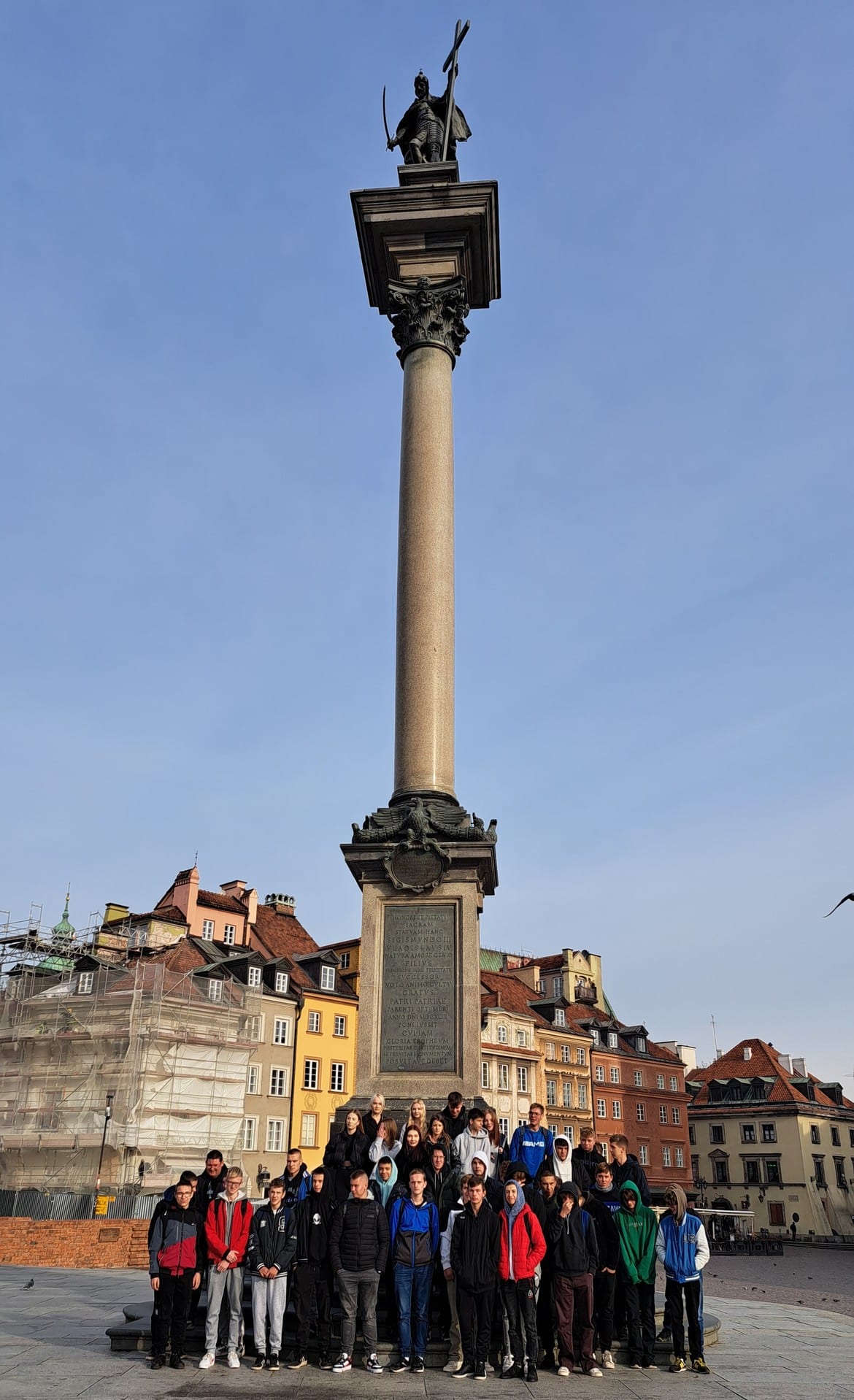 Grupa młodych ludzi stoi przed Kolumną Zygmunta w Warszawie, z widocznymi kamienicami w tle i błękitnym niebem.