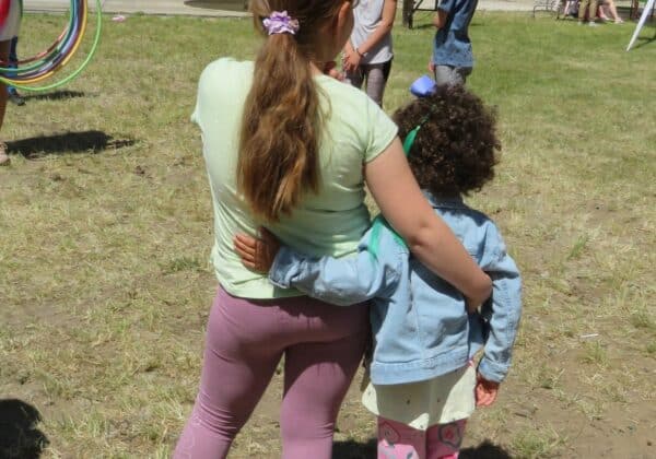 Na zdjęciu dwie dziewczynki przytulają się, obserwując grupę dzieci bawiących się na świeżym powietrzu podczas eventu.