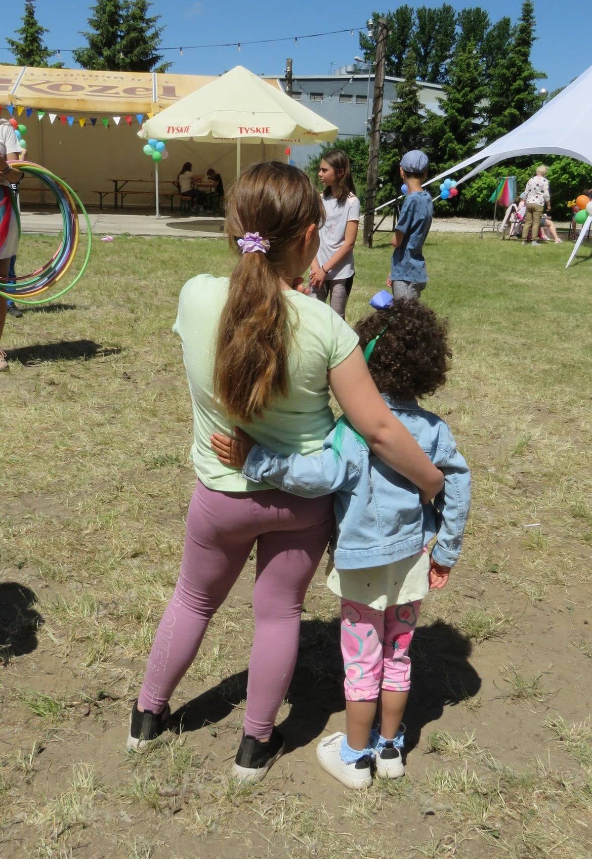 Na zdjęciu dwie dziewczynki przytulają się, obserwując grupę dzieci bawiących się na świeżym powietrzu podczas eventu.