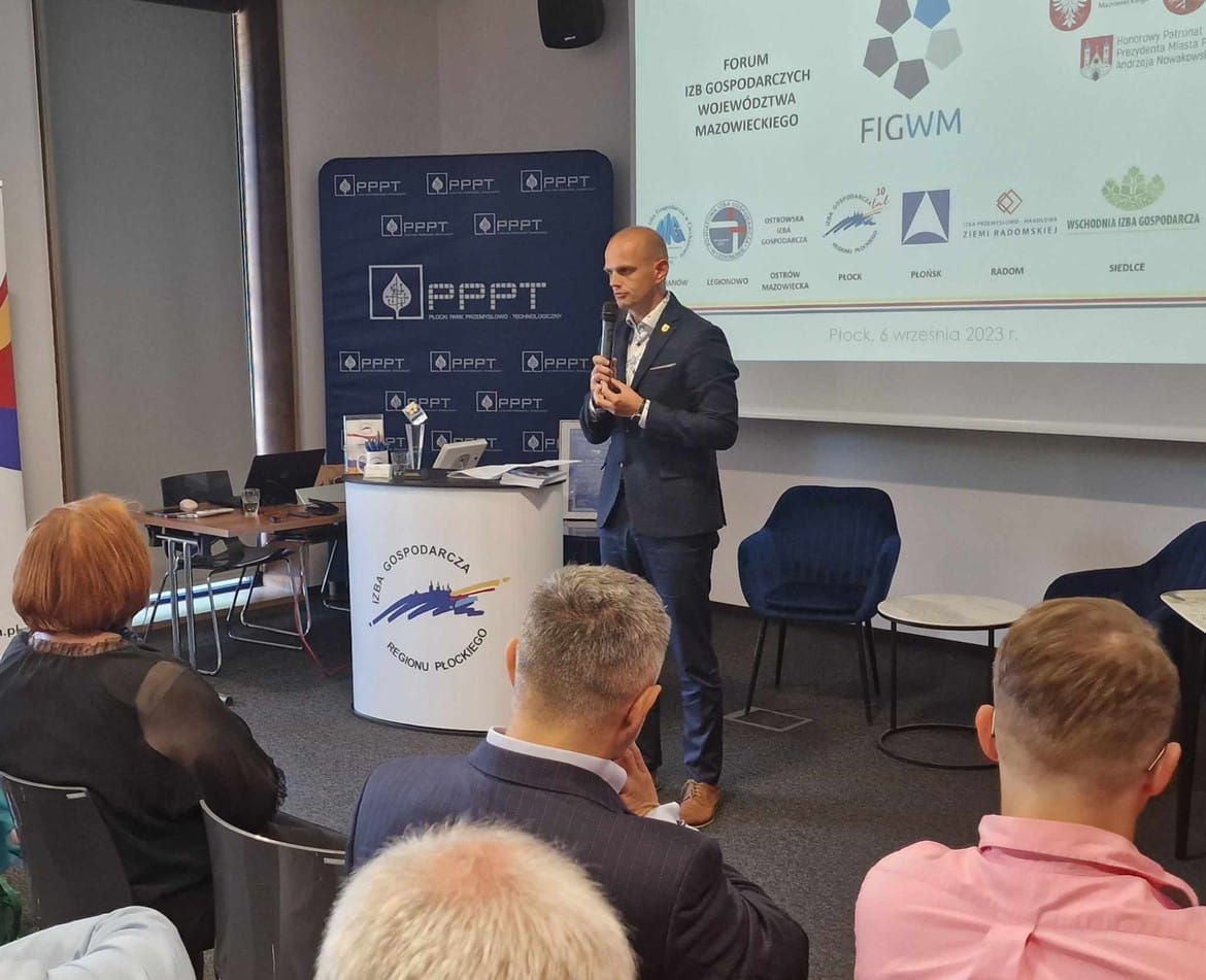 Zdjęcie przedstawia mężczyznę przemawiającego podczas spotkania w Izbie Gospodarczej Regionu Płockiego, z widokiem na uczestników konferencji.