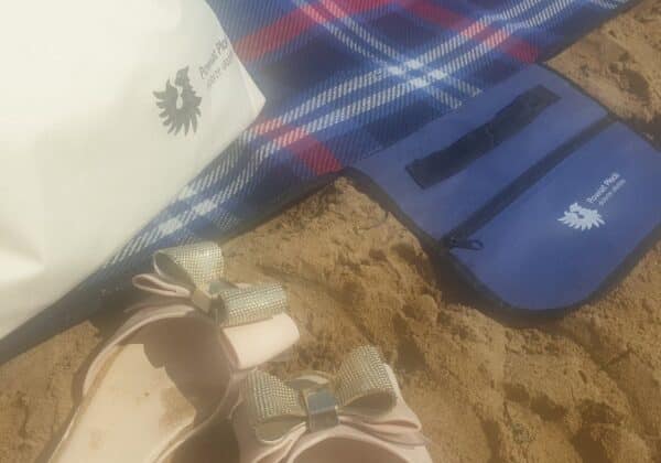 Na zdjęciu znajdują się różowe klapki z ozdobnymi kokardkami, biała torba oraz niebieska saszetka na plaży, na tle kolorowego koca.