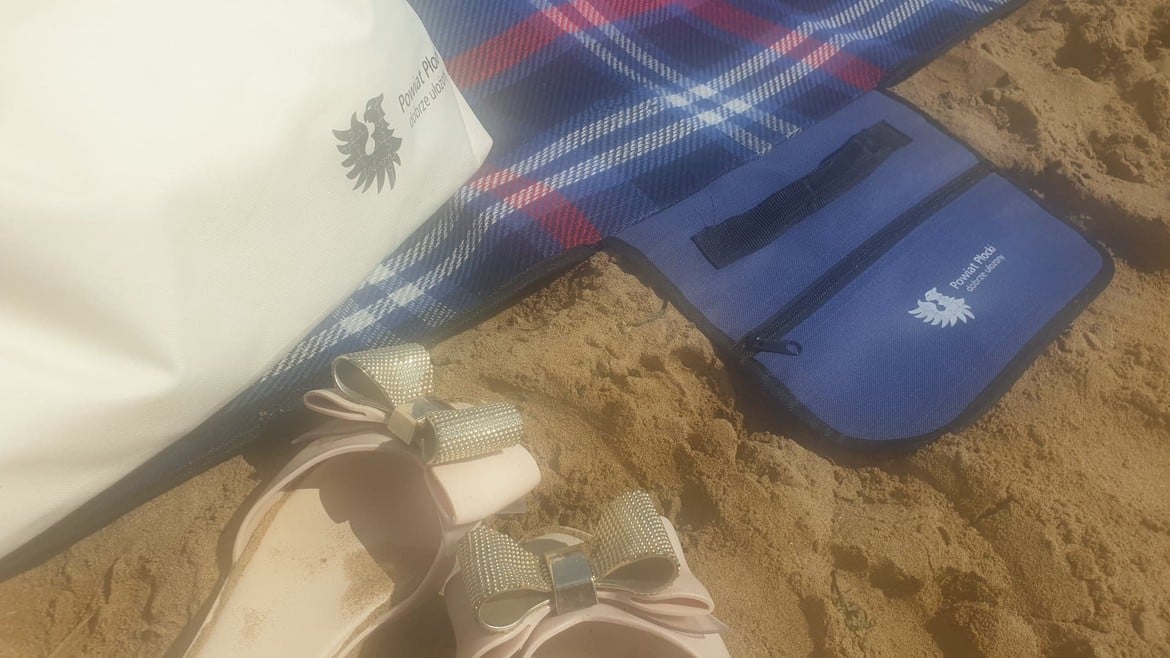 Na zdjęciu znajdują się różowe klapki z ozdobnymi kokardkami, biała torba oraz niebieska saszetka na plaży, na tle kolorowego koca.