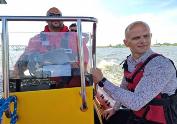 Na zdjęciu widać mężczyznę sterującego motorówką na wodzie oraz dwóch innych mężczyzn w tle, wszyscy w kamizelkach ratunkowych.