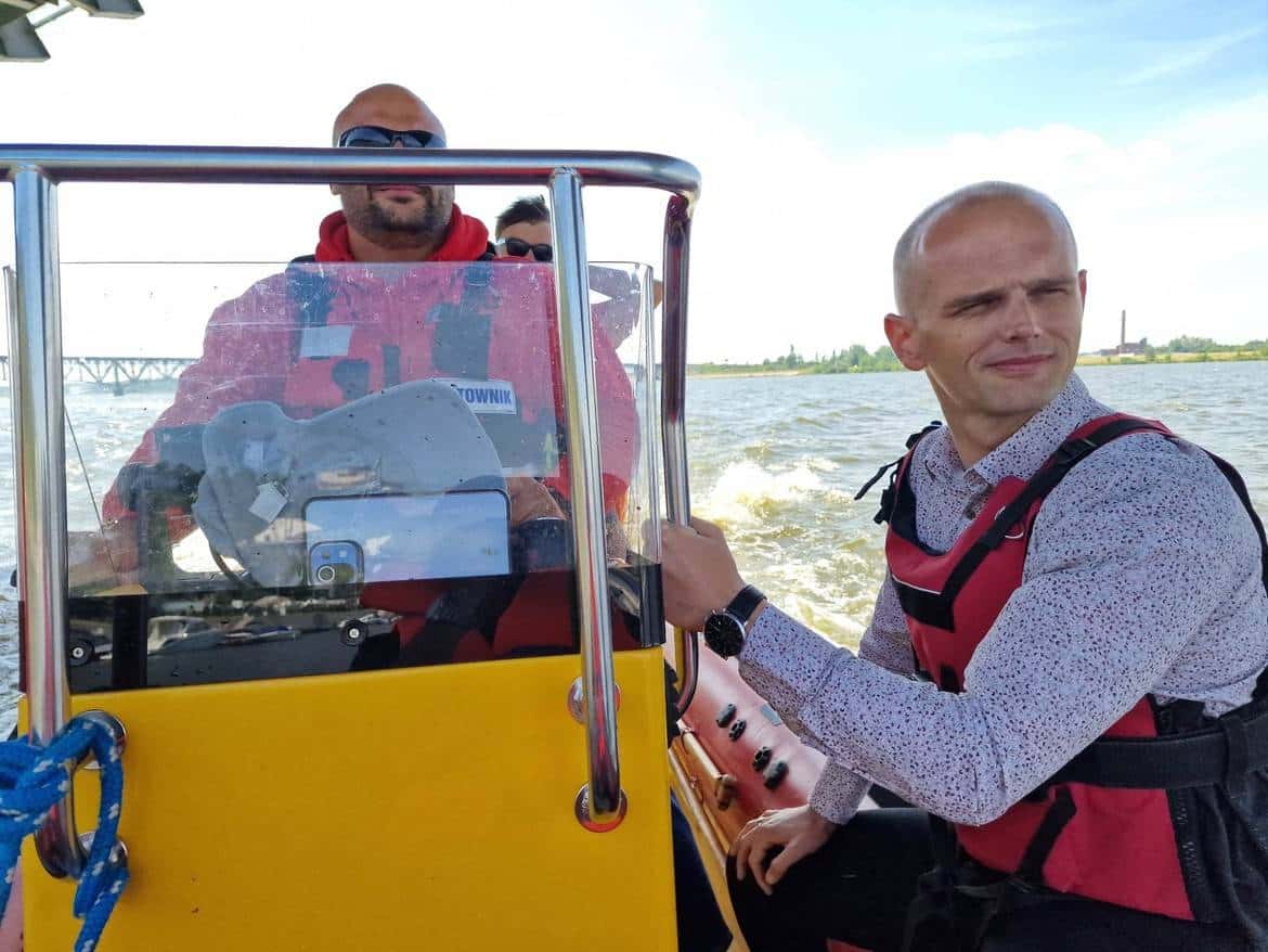 Na zdjęciu widać mężczyznę sterującego motorówką na wodzie oraz dwóch innych mężczyzn w tle, wszyscy w kamizelkach ratunkowych.