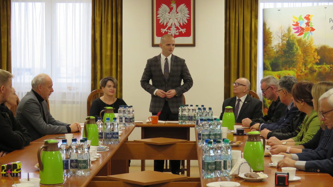 Na zdjęciu przedstawiono spotkanie w sali konferencyjnej, podczas którego mężczyzna w garniturze przemawia do zgromadzonych uczestników.