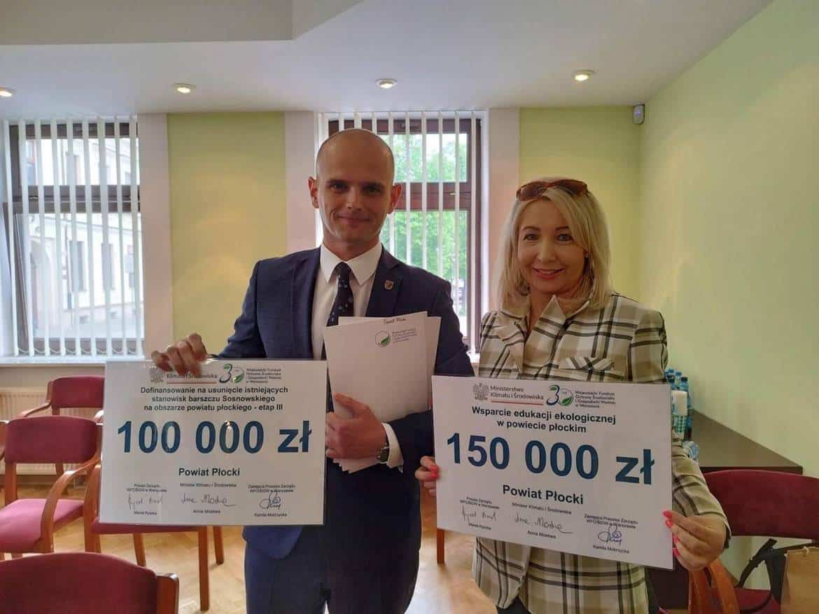Na zdjęciu dwie osoby trzymają plakaty informujące o przyznanych funduszach: 100 000 zł na usunięcie barczaka sosnowskiego i 150 000 zł na wsparcie edukacji ekologicznej w powiecie płockim.