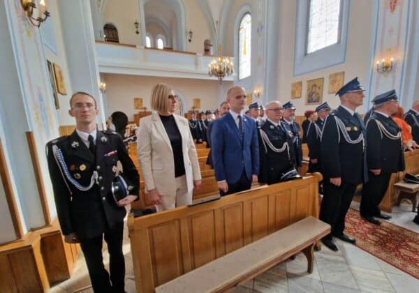 Uczestnicy ceremonii stoją w kościele, ubrani w mundury i eleganckie stroje, z poważnymi wyrazami twarzy.