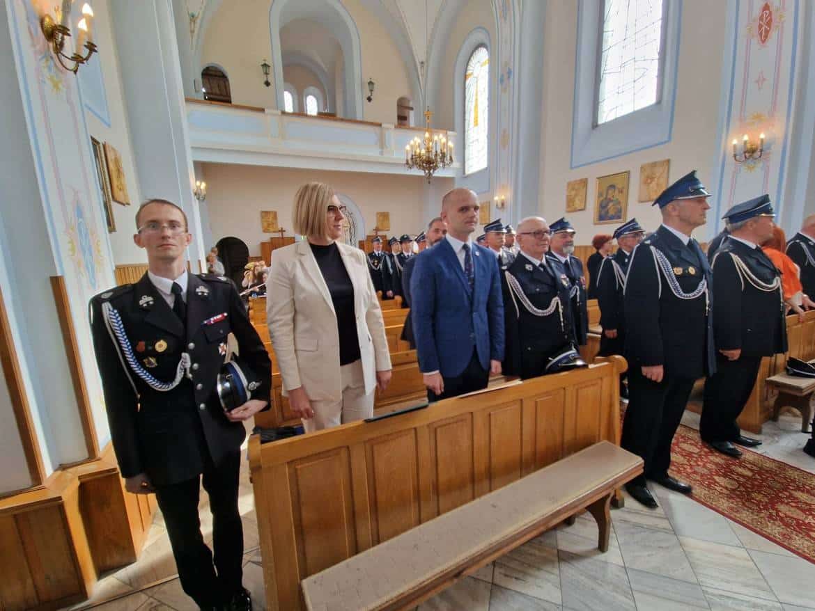 Uczestnicy ceremonii stoją w kościele, ubrani w mundury i eleganckie stroje, z poważnymi wyrazami twarzy.