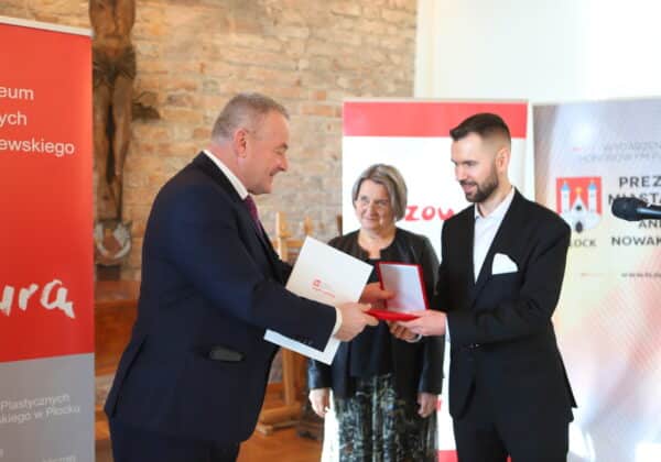 Na zdjęciu widać trzech ludzi w formalnych strojach, którzy uczestniczą w ceremonii wręczenia wyróżnienia, znajdując się w przestrzeni z kamiennymi ścianami i reklamami instytucji kulturalnych.