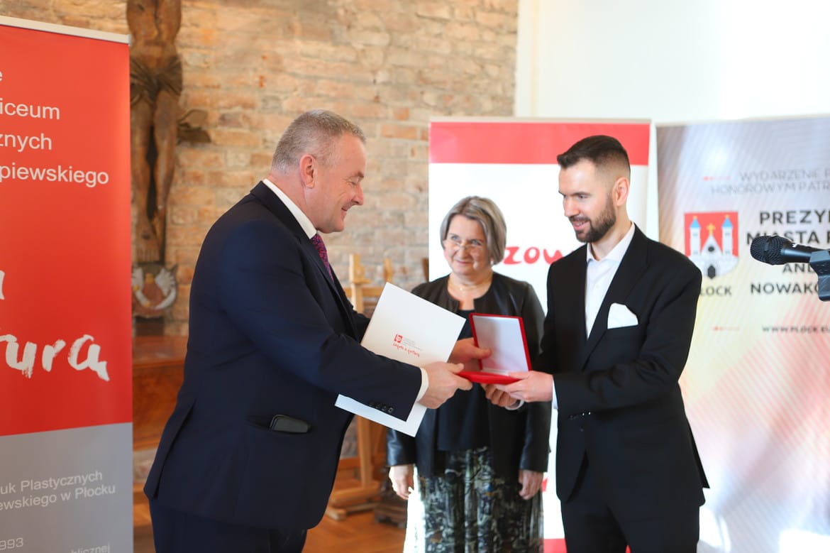 Na zdjęciu widać trzech ludzi w formalnych strojach, którzy uczestniczą w ceremonii wręczenia wyróżnienia, znajdując się w przestrzeni z kamiennymi ścianami i reklamami instytucji kulturalnych.