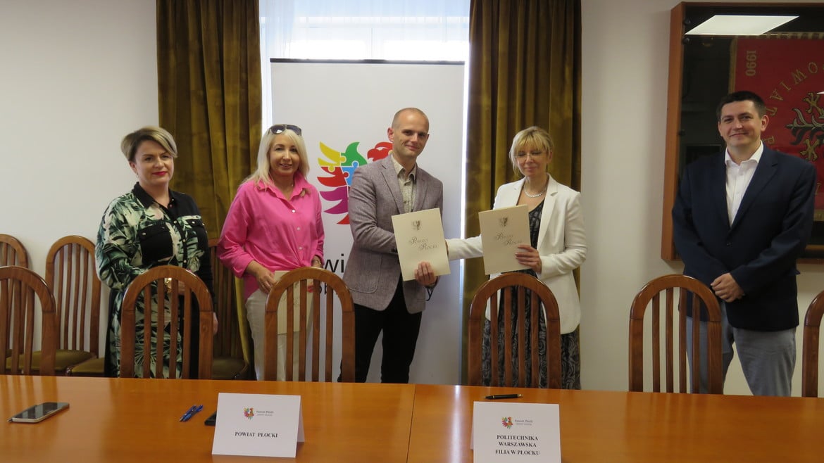Na zdjęciu przedstawiono grupę ludzi z podpisującymi dokumenty przy stole w sali spotkań, z logotypem Powiatu Płockiego w tle.