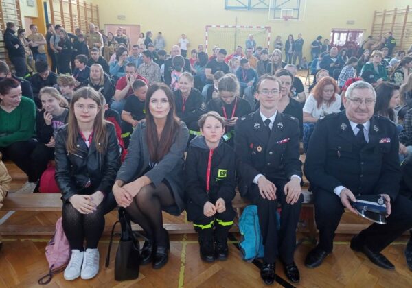 Grupa osób, w tym dzieci i dorosłych w mundurach, siedzi na ławkach w sali, otoczona przez tłum widzów.