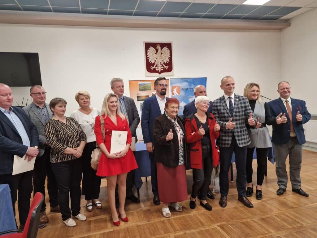 Zdjęcie przedstawia grupę ludzi z uśmiechami, trzymających kciuki w górę, na tle tablicy z logo oraz zdjęciami, podczas formalnego wydarzenia.