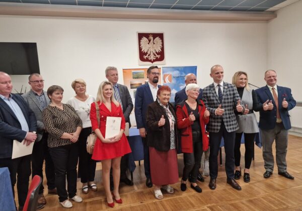 Na zdjęciu grupa ludzi stoi razem, uśmiechając się i pokazując gest kciuka w górę, w sali z dekoracjami w polskich barwach.