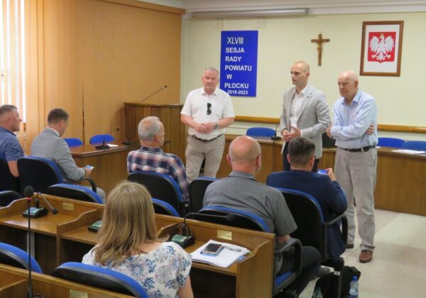 Na zdjęciu przedstawiono uczestników sesji rady powiatu w Płocku, gdzie kilku mężczyzn stoi na podium, a reszta słucha ich wystąpienia.