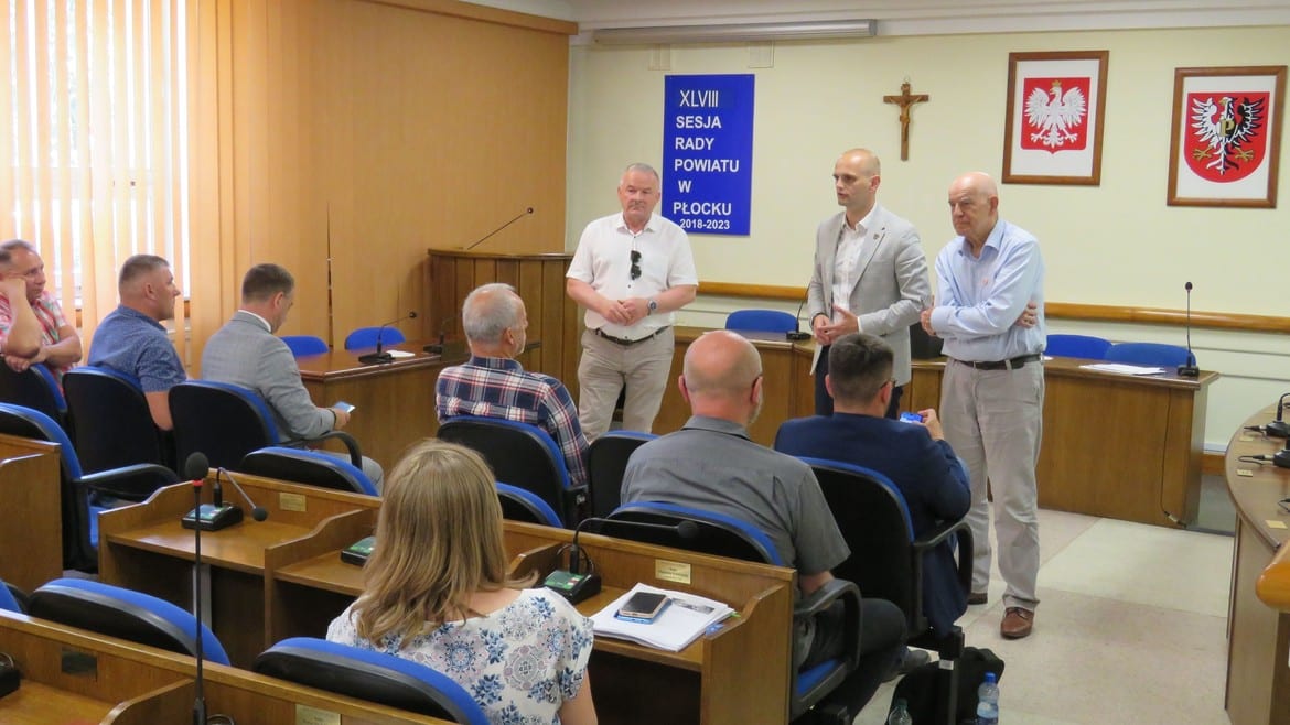 Na zdjęciu przedstawiono uczestników sesji rady powiatu w Płocku, gdzie kilku mężczyzn stoi na podium, a reszta słucha ich wystąpienia.