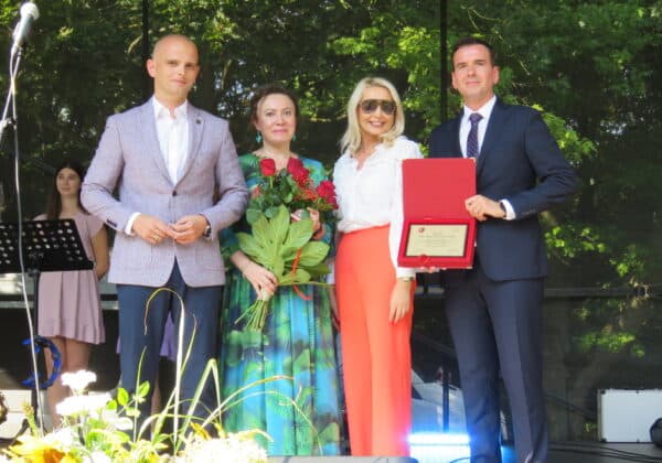Na zdjęciu widoczna jest grupa osób na scenie, która uczestniczy w ceremonii wręczenia dyplomu, a w tle znajdują się instrumenty muzyczne i zielone drzewa.