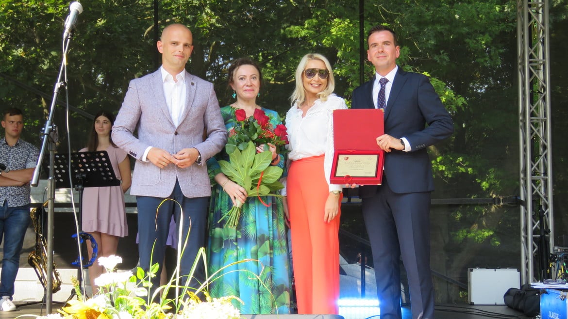 Na zdjęciu widoczna jest grupa osób na scenie, która uczestniczy w ceremonii wręczenia dyplomu, a w tle znajdują się instrumenty muzyczne i zielone drzewa.