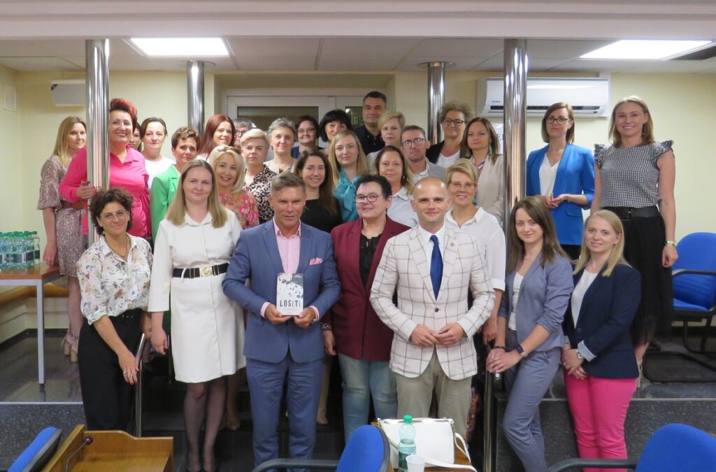 Grupa ludzi w formalnych strojach pozującą razem na tle sali, z jednym mężczyzną trzymającym książkę w centrum zdjęcia.