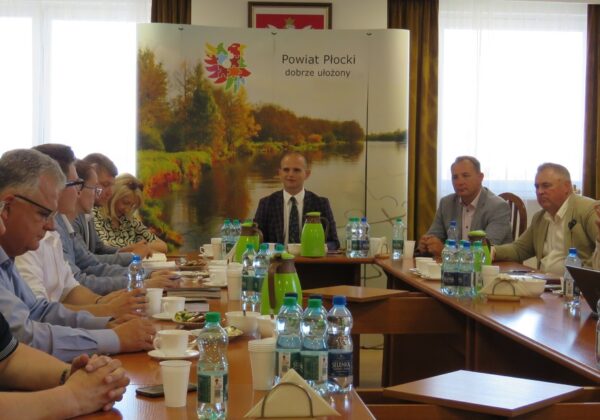 Na zdjęciu widać uczestników spotkania w sali konferencyjnej, którzy dyskutują przy długim stole, z tłem promującym Powiat Płocki.
