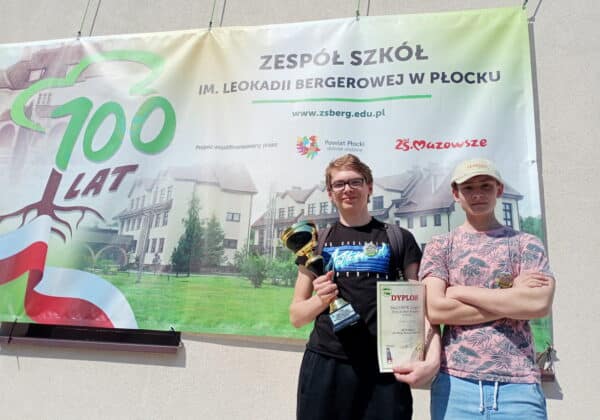Dwóch młodych mężczyzn stoi przed banerem z okazji 100-lecia Zespołu Szkół im. Leokadii Bergerowej w Płocku, trzymając trofea i dyplomy.