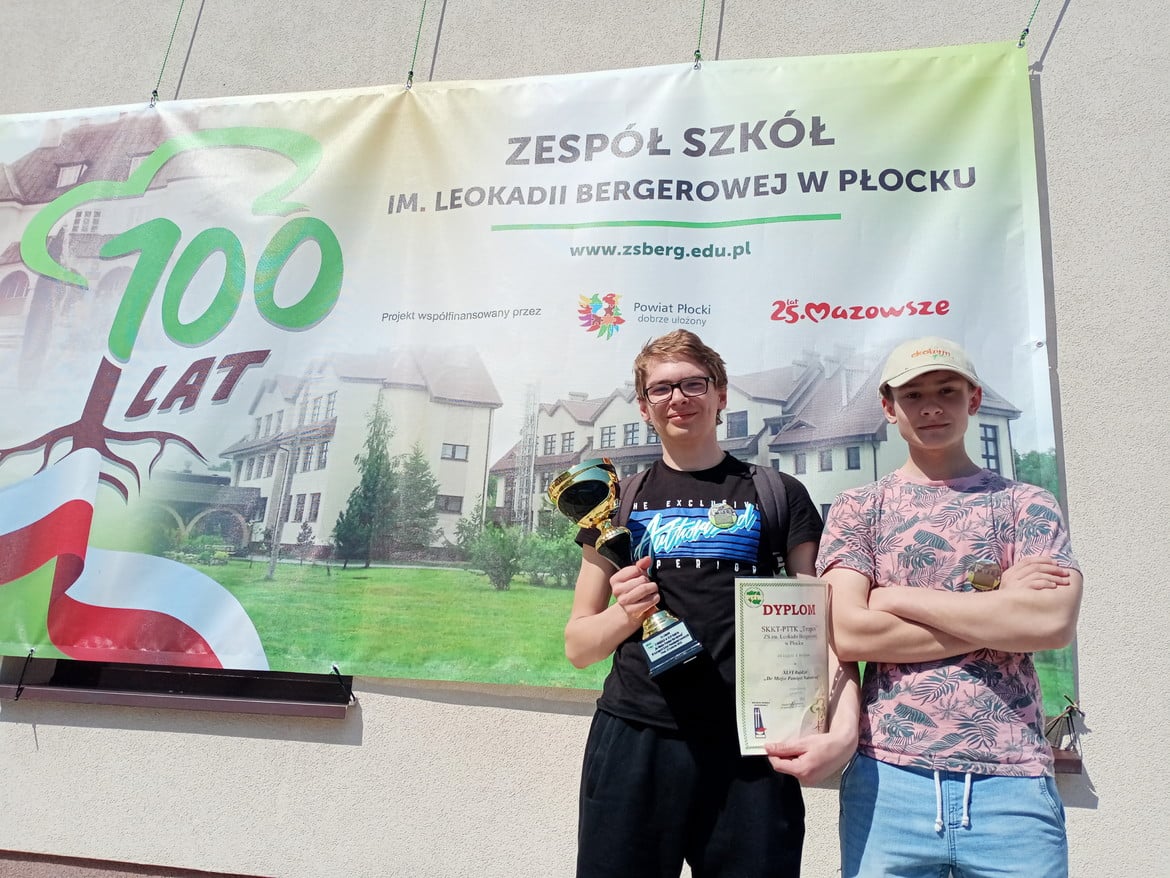 Dwóch młodych mężczyzn stoi przed banerem z okazji 100-lecia Zespołu Szkół im. Leokadii Bergerowej w Płocku, trzymając trofea i dyplomy.