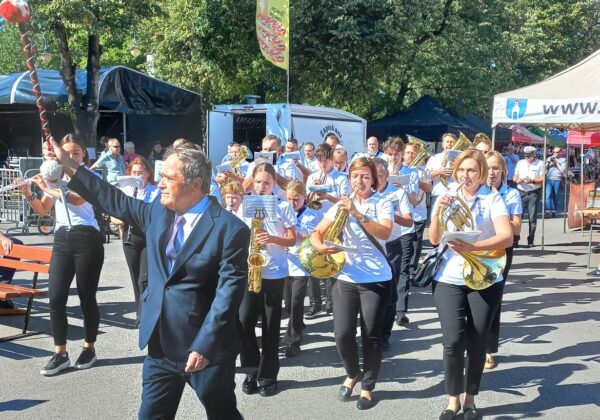 Na zdjęciu widać orkiestrę marszową, prowadzoną przez mężczyznę w garniturze, idącą w trakcie parady, w otoczeniu publiczności i straganów.