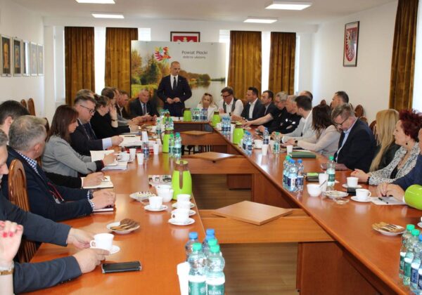 Na zdjęciu przedstawiono zebranie w sali konferencyjnej, z udziałem licznych uczestników za stołem, przy którym omawiane są różne tematy.