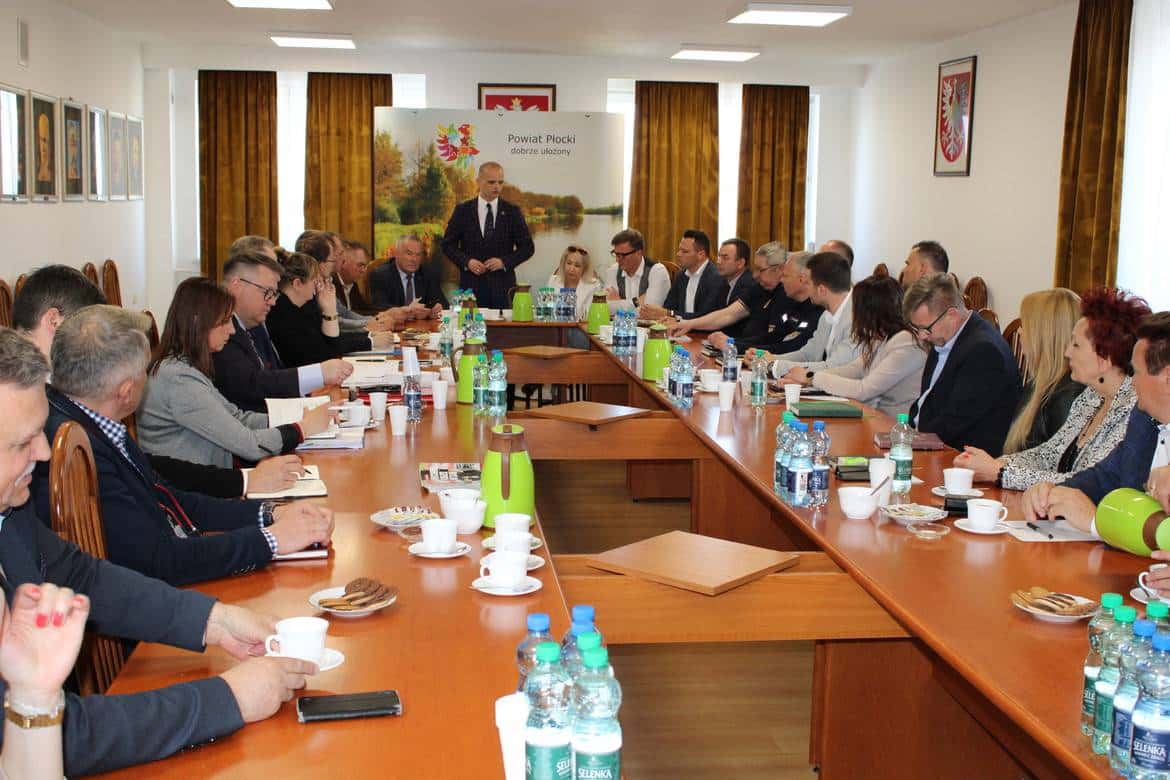 Na zdjęciu przedstawiono zebranie w sali konferencyjnej, z udziałem licznych uczestników za stołem, przy którym omawiane są różne tematy.