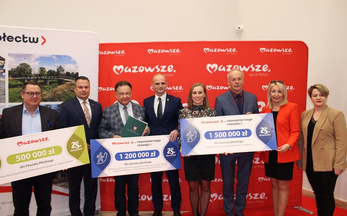 Na zdjęciu grupa osób stoi z dużymi czekami przedstawiającymi przyznane fundusze dla powiatu płockiego, w tle widoczne logo Mazowsza.