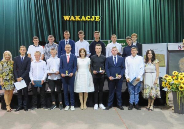 Grupa uczniów z nauczycielami stoi na scenie podczas uroczystości wręczenia nagród, w tle widoczne dekoracje i portret.