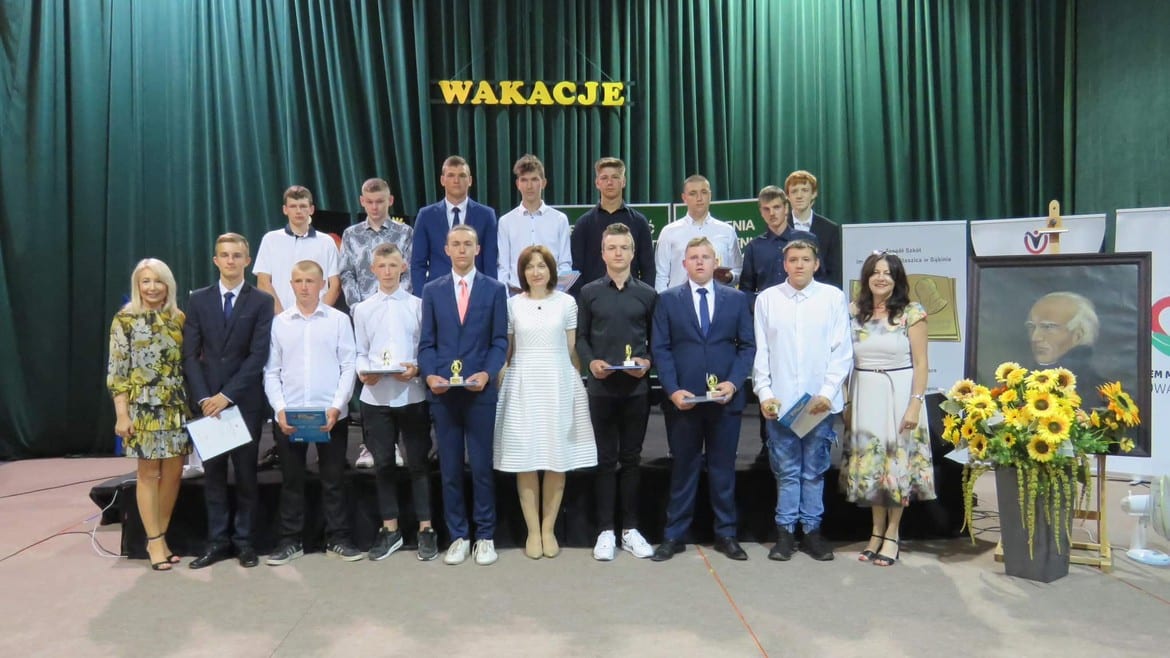 Grupa uczniów z nauczycielami stoi na scenie podczas uroczystości wręczenia nagród, w tle widoczne dekoracje i portret.