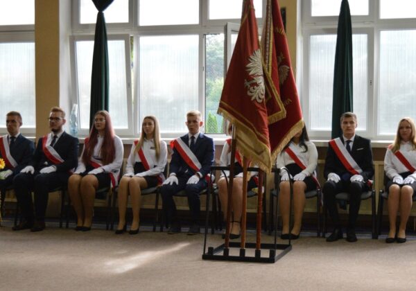 Grupa młodych ludzi w eleganckich strojach z odznakami siedzi w rzędzie, a obok nich stoją sztandary.